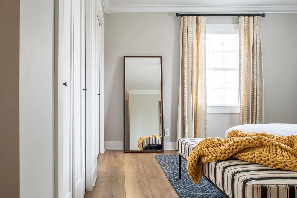 Image in a bedroom looking at a striped, upholstered bench with a yellow throw blanket and floor mirror in the background