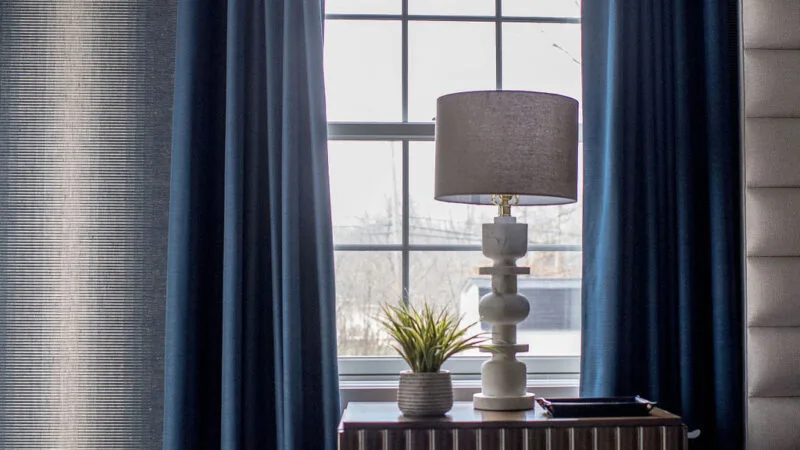 Table lamp sitting on top of a dark wooden nightstand in front of a window with blue panel drapes